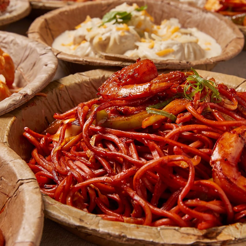Spicy red noodles with shrimp and vegetables in a bowl, with creamy momos in the background.